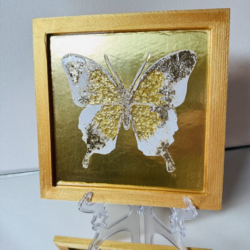 Ein strahlender Schmetterling aus strukturierter Paste auf goldenem Grund besticht durch goldenen Blütenstaub auf den Flügeln und einen schimmernden Kristall im Inneren. Der Schmetterling ist mit Epoxidharz überzogen, sodass er sich haptisch angenehm anfühlt.