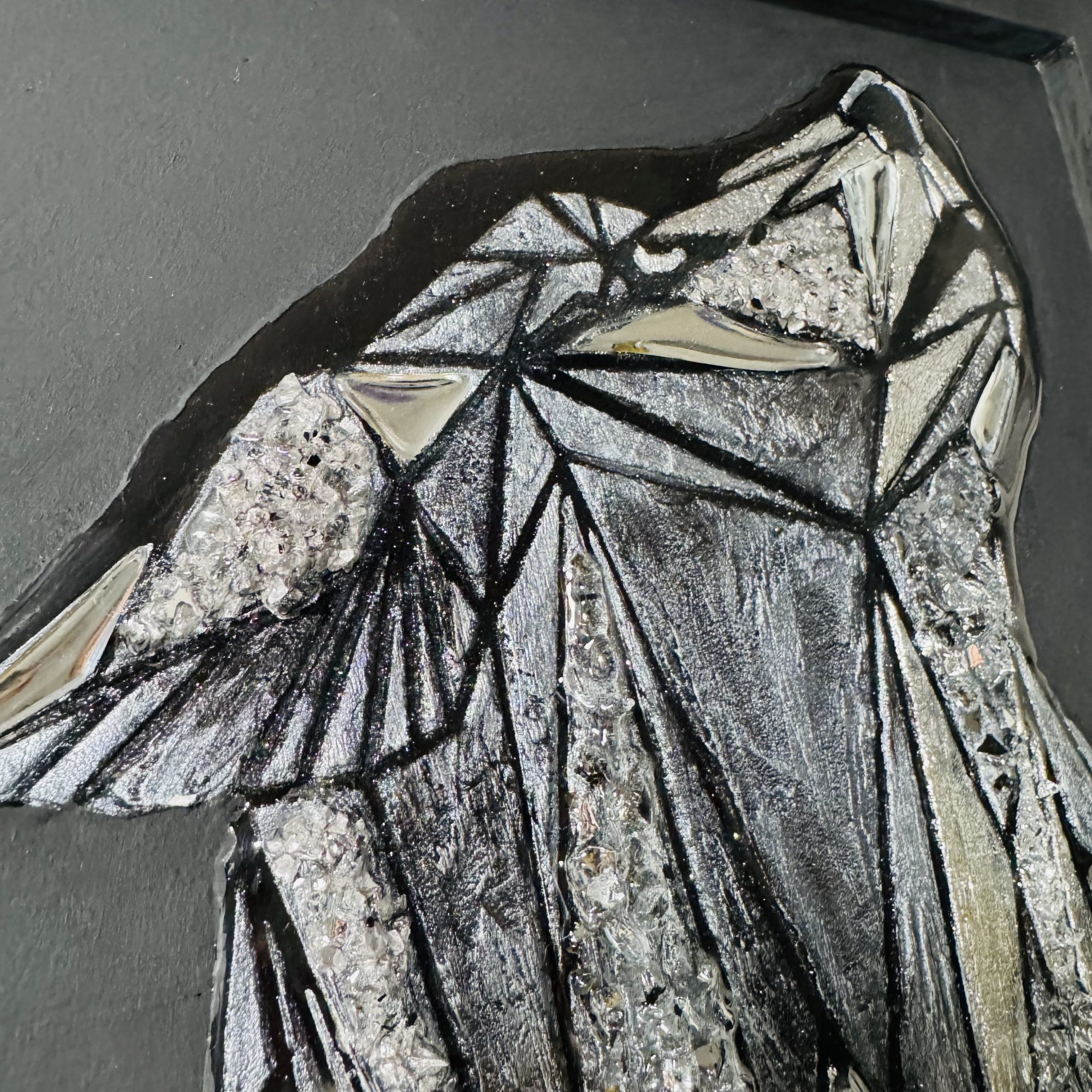 Ein grafisches, schimmerndes Bild eines Wolfskopfes auf mattschwarzem Hintergrund. Das Bild besteht aus handgeschnittenen, spiegelnden Dreieckselementen, strukturierter Paste, die mit schimmernder Farbe bemalt wurde, und spiegelnden Kristallen, die die Leuchtkraft des Gemäldes verstärken. Das Wolfsbild ist mit Epoxidharz überzogen, wodurch die zentrale Komposition mit dem Licht spielt und dem Betrachter aufregende Momente schenkt. Dadurch lässt sich das Gemälde auch berühren und haptisch erleben