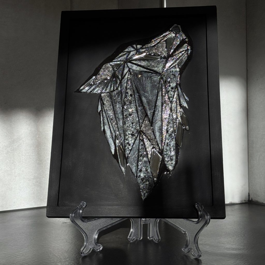 Ein grafisches, schimmerndes Bild eines Wolfskopfes auf mattschwarzem Hintergrund. Das Bild besteht aus handgeschnittenen, spiegelnden Dreieckselementen, strukturierter Paste, die mit schimmernder Farbe bemalt wurde, und spiegelnden Kristallen, die die Leuchtkraft des Gemäldes verstärken. Das Wolfsbild ist mit Epoxidharz überzogen, wodurch die zentrale Komposition mit dem Licht spielt und dem Betrachter aufregende Momente schenkt. Dadurch lässt sich das Gemälde auch berühren und haptisch erleben