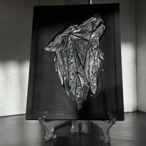 Ein grafisches, schimmerndes Bild eines Wolfskopfes auf mattschwarzem Hintergrund. Das Bild besteht aus handgeschnittenen, spiegelnden Dreieckselementen, strukturierter Paste, die mit schimmernder Farbe bemalt wurde, und spiegelnden Kristallen, die die Leuchtkraft des Gemäldes verstärken. Das Wolfsbild ist mit Epoxidharz überzogen, wodurch die zentrale Komposition mit dem Licht spielt und dem Betrachter aufregende Momente schenkt. Dadurch lässt sich das Gemälde auch berühren und haptisch erleben